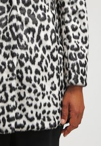 Abrigo de estampado de leopardo con una textura suave y peluda. La coloración en blanco y negro presenta manchas audaces. Mangas cortas y un corte recto.
