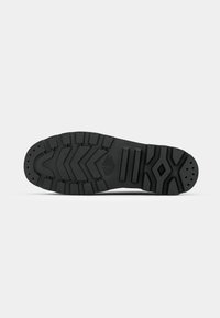 Semelle de botte en caoutchouc noir avec des motifs de bandes de roulement profondes, comprenant des chevrons, des rectangles et des formes en losange, sur un fond uni gris clair.