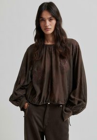 Blouse brodée marron avec un élastique à la taille, des manches longues en ballon et des motifs floraux dans un tissu léger et transparent.