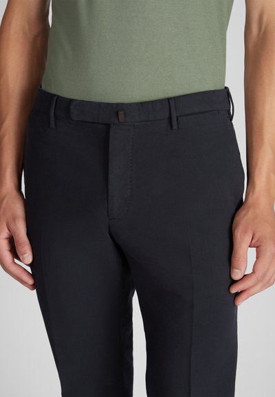 Close-up van een persoon die donker marineblauwe op maat gemaakte broek met riemlussen en een groene ingestopte shirt draagt, met de handen ontspannen aan de zijkanten.