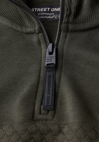 Olivově zelená bunda na zip s žebrovanou texturou, s černým zipem a štítkem "HLEDEJTE DOBRODRUŽSTVÍ", spolu s tkaným detailem na spodním lemu.