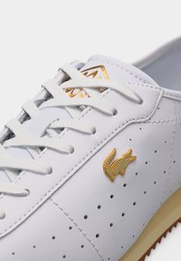 Λευκό δερμάτινο αθλητικό παπούτσι με λευκά κορδόνια, μικρές οπές στο πλάι και χρυσό λογότυπο κροκόδειλου κοντά στη φτέρνα.