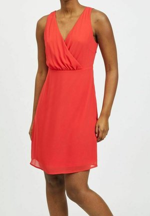 Woman wearing sleeveless red V-neck dress with wrap-style bodice, standing against plain light background.