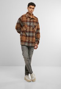 Bruine geruite flanellen shirt met twee voorzakken, knoopsluiting en lange mouwen, gecombineerd met versleten grijze jeans en witte sneakers.