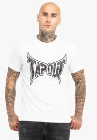 Balts kokvilnas T-krekls ar izteiksmīgu, nolietotu melnu "TAPOUT" logotipu priekšpusē. Īsas piedurknes un klasiskā apļa kakla dizains.