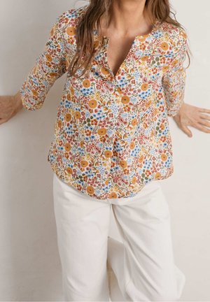 Kobieta w kwiecistej bluzce z długim rękawem z żółtymi, czerwonymi i niebieskimi kwiatami, w połączeniu z białymi, wysokim stanem spodniami, opierająca się o ścianę.