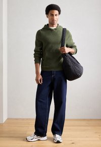 Felpa verde lavorata a maglia con cappuccio e tasca a marsupio, jeans blu a gamba larga, borsa trapuntata nera e sneakers bianche con dettagli grigi, su pavimento in legno.