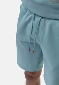 Pantalones cortos de color azul claro hechos de tela suave, con una cintura elástica y cuerdas ajustables con puntas rojas. Textura suave, diseño hasta la rodilla.