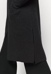 Černý pletený cardigan s bočním rozparkem, texturovanou tkaninou a přední kapsou, doplněný černými legínami.