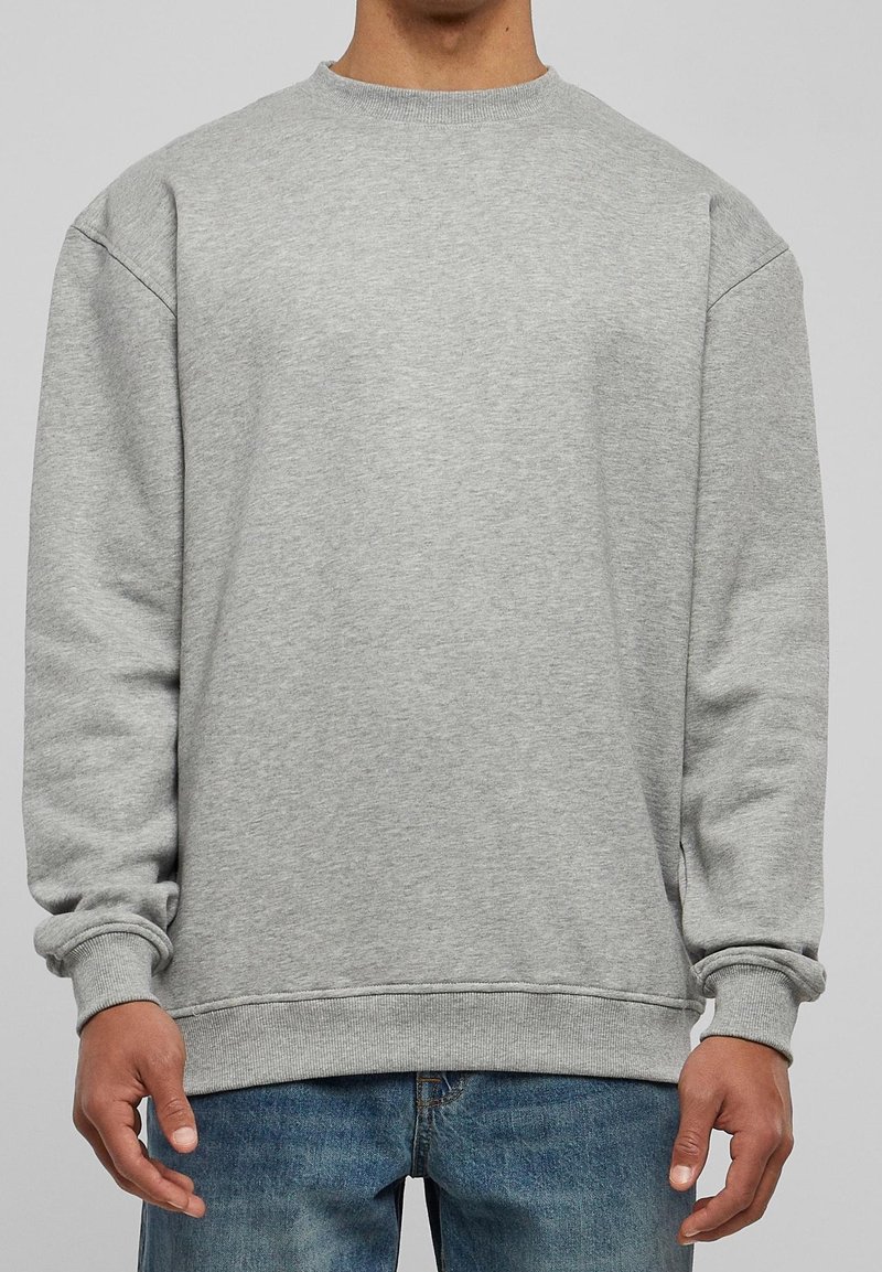Personne portant un sweat-shirt gris clair uni à col rond avec des manches longues et un jean bleu, debout devant un fond neutre.