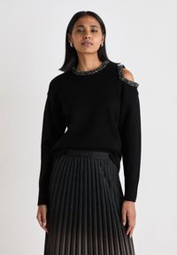 Μαύρος πουλόβερ με σχέδιο κρύου ώμου και κεντημένη λαιμόκοψη, συνδυασμένο με πλισέ μαύρη φούστα που διαθέτει μεταλλική γυαλάδα και ελαστική μέση.