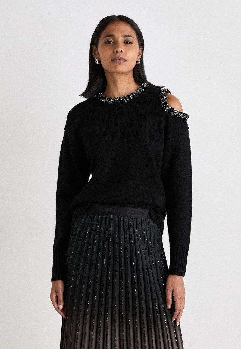 Μαύρος πουλόβερ με σχέδιο κρύου ώμου και κεντημένη λαιμόκοψη, συνδυασμένο με πλισέ μαύρη φούστα που διαθέτει μεταλλική γυαλάδα και ελαστική μέση.