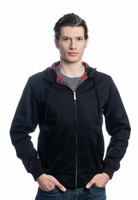 Veste à capuche zippée noire avec doublure en tartan rouge, poches latérales, poignets côtelés et finition texturée. Portée par-dessus une chemise grise.