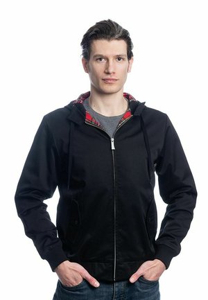 Veste à capuche zippée noire avec doublure en tartan rouge, poches latérales, poignets côtelés et finition texturée. Portée par-dessus une chemise grise.