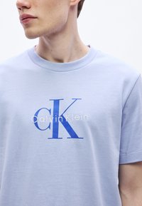 Hele sinine puuvillane t-särk ümarate kaelustega, millel on sinise ja valge graafikuga tekst "Calvin Klein" ja "CK". Sile tekstuur.