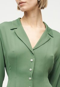 Γυναίκα που φοράει στενή πράσινη πουκάμισο με κουμπιά, γιακά με εγκοπή και μακριά μανίκια, εμφανιζόμενη από τον λαιμό μέχρι τη μέση.