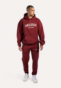 Bordeaux hoodie met een voorkwab en witte tekst, gecombineerd met bijpassende bordeaux sweatpants. Heeft ribbelmanchetten en witte sneakers.