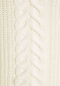 Suéter blanco de punto que presenta un diseño de trenzas prominente con patrones texturizados y costuras abiertas, destacando un hilo grueso y una intrincada artesanía.