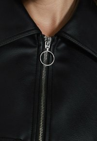 Collier de veste en cuir synthétique noir avec une fermeture éclair en métal à l'avant et une tirette circulaire. Surface texturée et détails de coutures visibles.