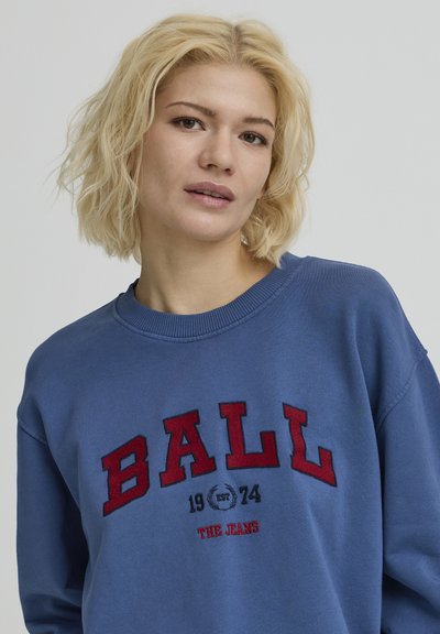 Sweat-shirt bleu avec un col rond et une broderie rouge texturée. Le devant affiche en lettres grasses "BALL 1974 THE JEANS".