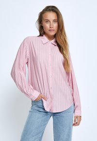 Camicia a maniche lunghe rosa chiaro con bottoni e strisce verticali bianche, dotata di colletto aperto e orlo arrotondato, abbinata a jeans blu chiaro.