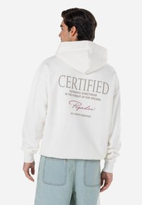 Biela mikina s kapucňou a veľkým nápisom "CERTIFIED" na chrbte. Obsahuje šnúrku na kapucni, rebrované manžety a voľný strih. Skombinovaná s svetlomodrými nohavicami.