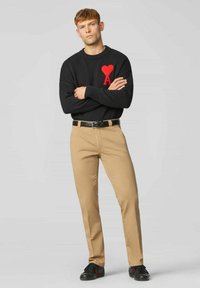 Zwarte trui met een rood hart en een "A"-grafiek, beige broek, zwarte sneakers met rode accenten, staand met de armen gekruist tegen een neutrale achtergrond.