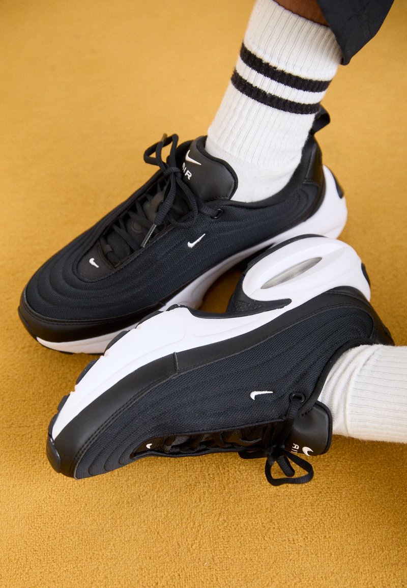 Pieds portant des chaussures Nike Air Max noires et blanches avec des chaussettes blanches à deux bandes noires, debout sur une surface jaune.