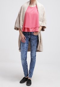 Haut à plusieurs couches rose clair, manteau beige oversize, jean skinny bleu avec accents piqués, et chaussures noires. Textures douces et design décontracté.