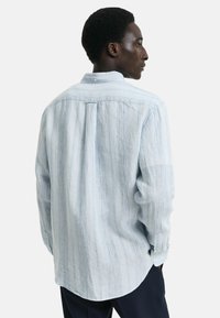 Camisa a rayas azul claro con una textura suave, con cuello abotonado y mangas largas. Presenta un dobladillo curvado y detalles con pliegues en la parte posterior.