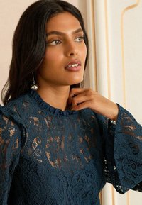 Mornarsko plava čipkasta majica s volanima oko ovratnika, širokim zvonastim rukavima i složenim cvjetnim uzorcima. Tkanina ima delikatnu teksturu.