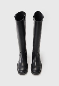 Bottes noires en cuir, montantes jusqu'au genou, avec une texture lisse, un bout rond et un minimalisme au niveau des coutures, dotées d'une languette en haut.
