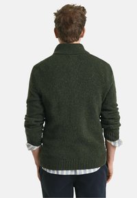 Dunkelgrüner gestrickter Pullover mit hohem Kragen, gerippten Bündchen und Saum. Über einem hellblauen und weißen gestreiften Hemd getragen. Tolle Texturdetails.
