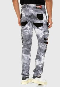 Homme portant un pantalon cargo tie-dye gris et blanc avec des accents de poches noires et des baskets blanches à plateforme, se tenant de dos.