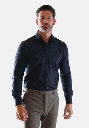 Man with dark hair and beard wearing a fitted dark blue button-up shirt and beige dress pants against a plain light background.