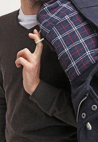 Mörkbrun tröja, vit t-shirt under, håller en marinblå kappa med rutigt foder som har röda och vita rutmönster.