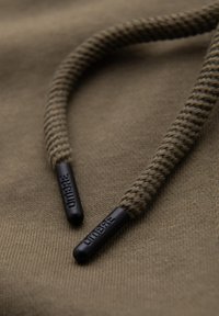 Gros plan sur des cordons de capuche tressés marron avec embouts en plastique noirs embossés "OMBRÉ" sur un tissu assorti.