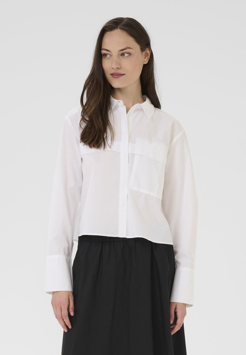 Bela srajca z dolgimi rokavi, dvema sprednjima žepoma in skrajšanim dizajnom. V kombinaciji z črno, tekočo krilom. Enostavna tekstura.