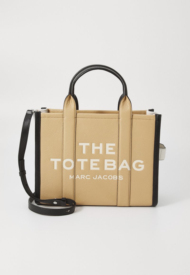 Tan leather tote bag with black accents, featuring dual top handles, an adjustable shoulder strap, and large white text: "THE TOTE BAG MARC JACOBS."