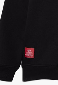 Sudadera negra con puños acanalados, con una etiqueta de marca roja que muestra texto y un logo en la parte inferior izquierda. Textura textil suave.