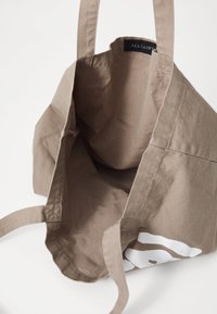Leichte braune Stofftasche mit zwei stabilen Schultergurten. Das Innere ist ungefüttert und auf einer Seite befindet sich ein einfaches weißes Grafikdesign.