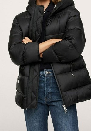 Person trägt schwarze gesteppte Winterjacke mit Kapuze und blaue Jeans, verschränkte Arme, steht vor einem schlichten hellen Hintergrund.