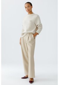 Hellbeige Pullover mit strukturiertem Finish, kombiniert mit hochgeschnittenen beigen Hosen. Burgunderfarbene spitz zulaufende Flats runden das Outfit ab.