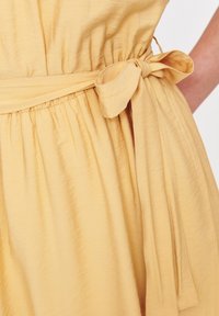 Żółta sukienka z wiązanym paskiem w talii, z marszczonymi teksturami i lekkim materiałem. Projekt obejmuje płynący materiał z delikatnym drapowaniem.