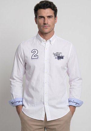Hombre con camisa blanca con botones y texto bordado en azul y número "2", de pie con las manos en los bolsillos contra un fondo neutro.