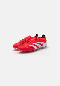 Red soccer cleats with a textured surface, featuring white stripes and a sleek design. Studded outsole for traction on grass.