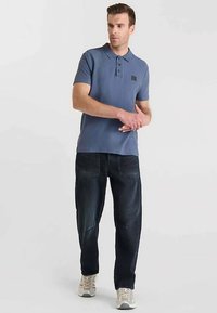 Polo bleu avec patte de boutonnage à trois boutons, associé à un jean sombre ample, porté avec des baskets de couleur claire. Marque discrète.