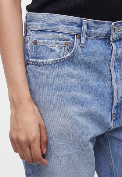 Το αριστερό χέρι ακουμπά δίπλα σε σκισμένα ανοιχτό μπλε τζιν παντελόνια, που φοριούνται με μαύρο τοπ, με ορατές λεπτομέρειες μέσης και τσέπης.