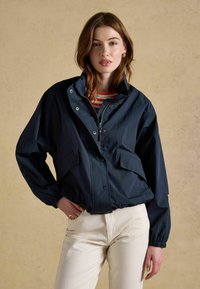 Młoda kobieta z rozpuszczonymi brązowymi włosami nosi granatową kurtkę na zamek, pod którą ma czerwono-białą koszulkę w paski oraz kremowe spodnie.