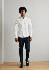 Hvit skjorte med knapper, mørke stripete bukser og svarte sko. Glatt stoff, tettsittende design og ensfargede farger. Stående posering mot en ensfarget bakgrunn.